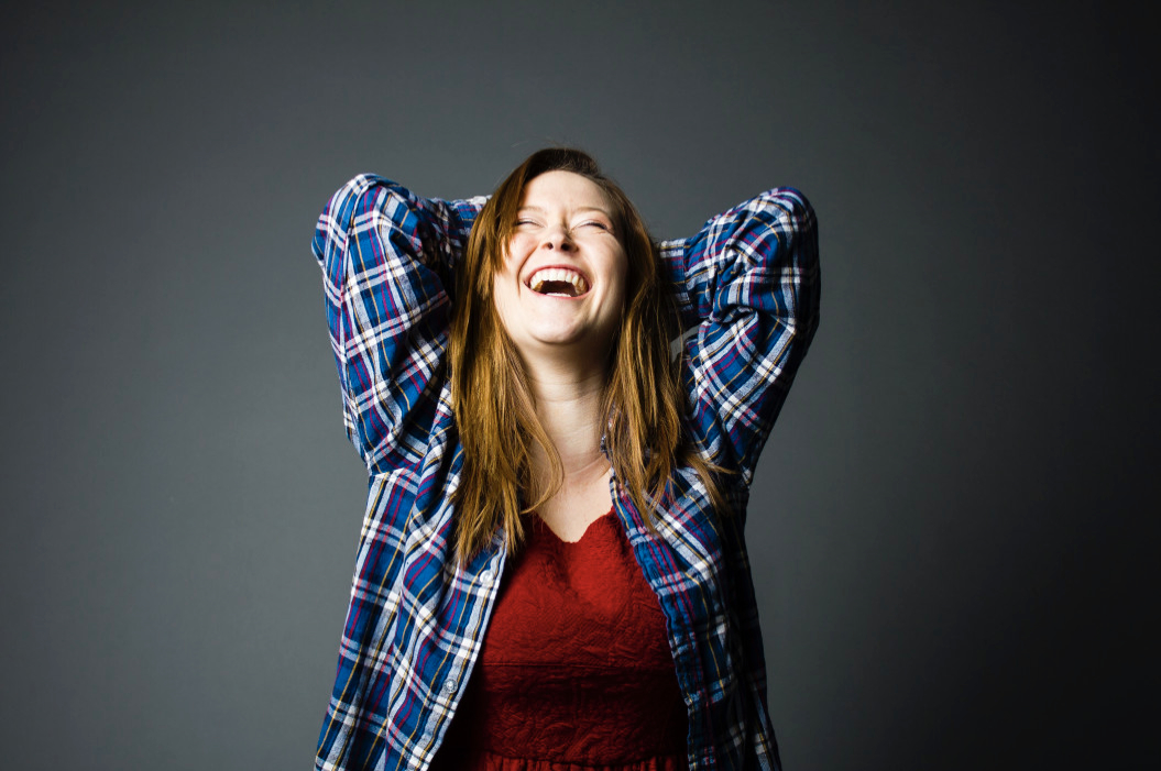 Woman on gray background, hands behind head, laughing